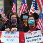 Asian women wear masks and hold up signs "we are Americans too!" and "STOP Asian Hate" with American flags behind them.