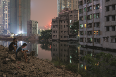two men sit on a pile of rubble by a river at night, with crumbling buildings, lights and new construction in the background