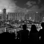 Beauty of the City by Timothy Tse. Black and white photo, three shadows sitting in the foreground looking out from atop a hill at a city of skyscrapers, lit up at night.