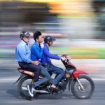 Three people ride a motorcycle, with a blurred background, in Vietnam. Photo by Timothy Tse.