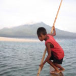 Boy holding a stick on a boat in the ocean and a cloudy mountain