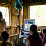 kids surround a karaoke machine in a small hut