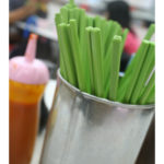 A container of green chopsticks and a bottle of hot sauce sit in the foreground, with blurry people eating at a table in the background.