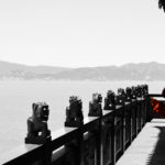 view of a fence with statues on the edge of a body of water with mountains in the distance.