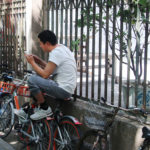 A man sits atop a bike leaning against a fence, reading on his phone.