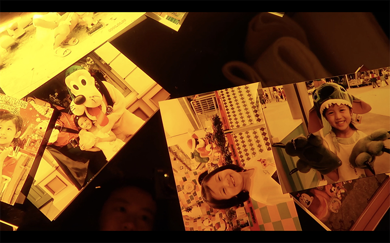 A still image from Cheryl Cheung's video "Being Western." Cast in a warm yellow hue, the image shows several photographs of a young child. Especially visible is an image of the child with a figure in a Pluto the dog costume.