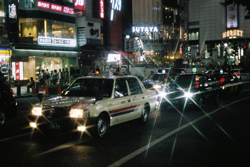 Photo in Andy Takagi’s “Summertime in Tokyo” series. the image shows Taxi traveling through Shibuya crossing at night