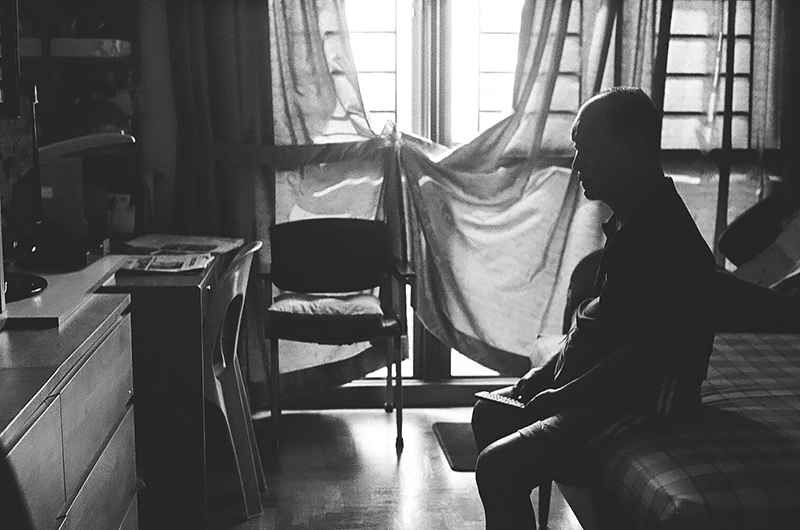 The first photo in Amanda Ann-Min Wong's series, Lest We Remember. The black and white photo shows an interior with a seated figure silhouetted in front of a curtained window.