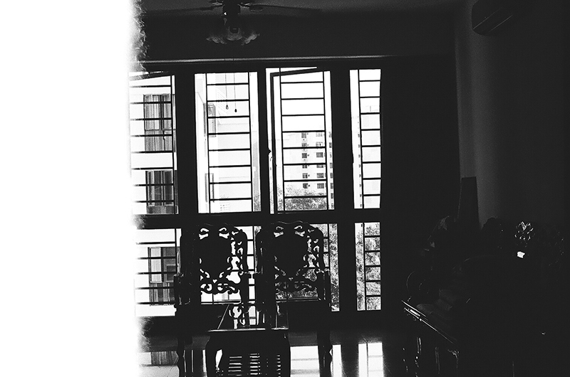 The second photo in Amanda Ann-Min Wong's photo series, Lest We Remember. The partially exposed black and white photo shows two ornate chairs silhouetted in front of a window.