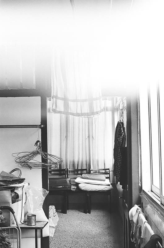 The third photo in Amanda Ann-Min Wong's series, Lest We Remember. A partially exposed black and white photo, the image shows the lower corner of a room with a small accumulation of objects such as clothes hangers and hanging clothing.