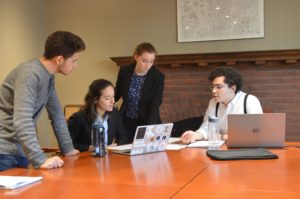 A group of 4 Munk One students stand around a table with chairs discussing ideas for their case competition.
