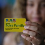 A young woman holds a Bolsa Familia membership card to the foreground.