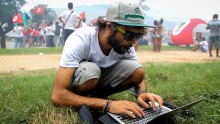 A man crouches in the grass with a laptop computer.