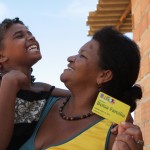 A woman holds a Bolsa Familia benefit card in one hand, and her child in the other
