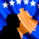A man stands in front of a screen displaying the Kosovo flag
