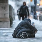 A homeless person sits in the street wrapped in a blanket