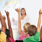A group of children in a classroom raise their hands