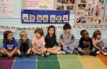 A group of pre-kindergarten students sit on the floor in their classroom