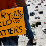 Child holding "every child matters" sign with shoes in the background on the cement