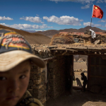 Tibetan Child in front of a stone building with a Chinese flag flying in the background